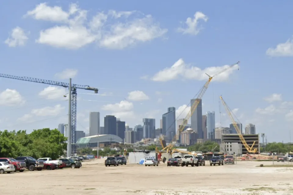 Dirt lot with construction cranes and parked cars with downtown Houston skyline in background